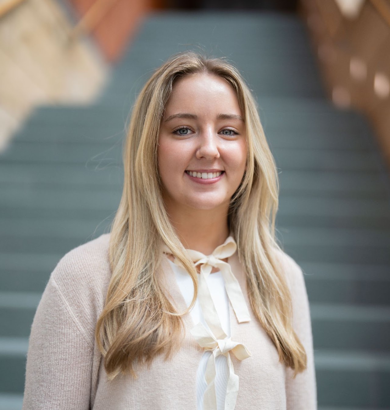 A professional headshot of Vaiva Raisys, a young woman with long blonde hair, smiling softly. She is wearing a light-colored sweater with a bow, in front of a blurred background of stairs.